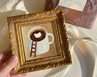 May include: A framed painting of a white coffee mug filled with dark liquid and a white duck. A small wooden ladder leads into the mug. The frame is gold with ornate detailing. A gift wrapped in brown paper with a tag that says "Thank You!" is in the background.