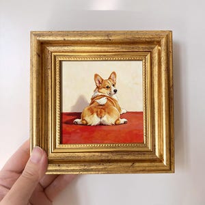 May include: A framed painting featuring a tan and white Corgi dog lying on a red surface, looking back over its shoulder. A small white bird is perched on the dog's back. The artwork is enclosed in a decorative, ornate gold-colored frame.