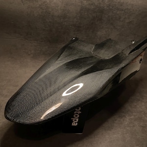 Może przedstawiać: Błyszczący błotnik motocyklowy z włókna węglowego o wyraźnej tkanej fakturze i gładkim, aerodynamicznym kształcie. Element ma ciemne, odblaskowe wykończenie i zawiera małą czarną metkę z częściowo widocznym białym napisem 'otopa'.