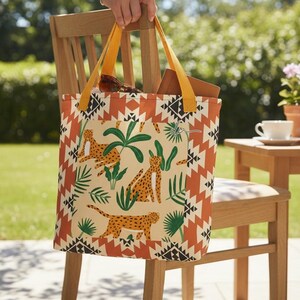 May include: A beige tote bag with orange and black geometric patterns and yellow straps. The bag features a repeating pattern of cheetahs and tropical plants. The bag is held up by a person.