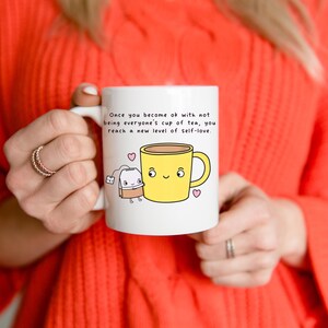 Puede incluir: Taza de cerámica blanca con una taza de té amarilla de dibujos animados y un personaje de bolsita de té, con el texto: "Once you become ok with not being everyone's cup of tea, you reach a new level of self-love."