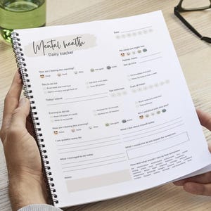 Puede incluir: Un rastreador diario de salud mental encuadernado en espiral con el título "Mental health" en un estilo manuscrito. El rastreador incluye secciones para el estado de ánimo, las tareas diarias y las notas. Las páginas son blancas con texto negro y acentos grises claros.