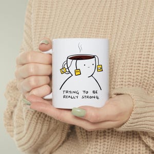 Puede incluir: Taza de cerámica blanca con una ilustración humorística de una taza de té con bolsitas de té y el texto "TRYING TO BE REALLY STRONG." La taza tiene un diseño de contorno negro.