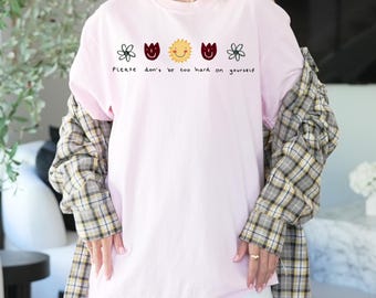Può includere: Una maglietta rosa chiaro con la scritta "Please don't be too hard on yourself" e grafiche di fiori e sole. La maglietta è realizzata in un materiale morbido.