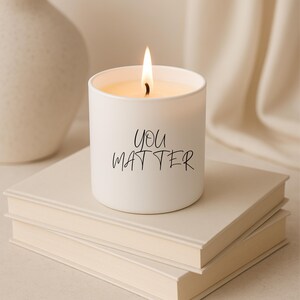 May include: A lit white candle with the words "You Matter" in black script. The candle sits atop two off-white books. A white vase is in the background, with a cream-colored fabric draped behind the candle.