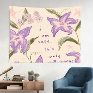 May include: A tapestry featuring a hand-drawn floral design with purple flowers, green leaves, and the text "i am safe, it's only change." The background is a light beige color, and the overall aesthetic is soft and artistic.