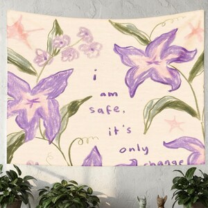 May include: A wall tapestry featuring a hand-drawn floral design with large purple flowers and green leaves. The text "I am safe, it's only change" is written in a handwritten font. The background is a cream color.