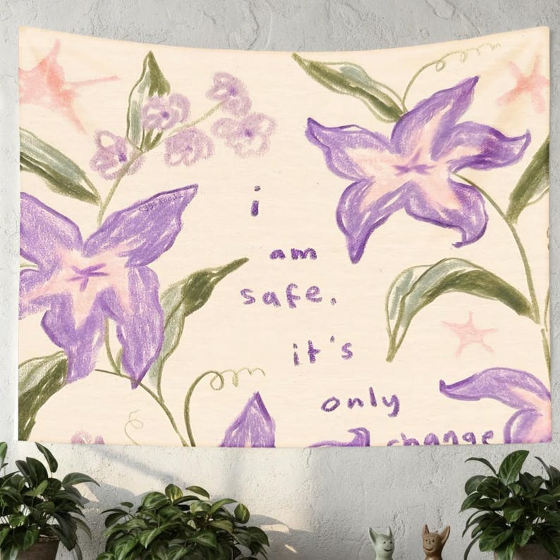 May include: A wall tapestry featuring a hand-drawn floral design with large purple flowers and green leaves. The text "I am safe, it's only change" is written in a handwritten font. The background is a cream color.