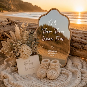 May include: A scalloped-edge mirror with the white text And Then There Were Four, a heart icon, and ARRIVING NOVEMBER 2026. Beside it sits a bouquet of dried flowers, a card reading Baby Henry, and a pair of beige knit baby booties on a cream blanket.