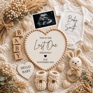 Peut inclure: Un body blanc avec le texte Baby Smith, un panneau en forme de cœur indiquant This is the Last One Seriously! Due November 2026, une échographie, des cubes en bois, des chaussons tricotés, un hochet lapin et des fleurs séchées sur un tissu crème.