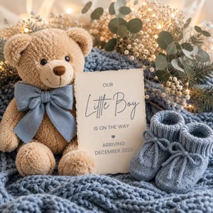 Pode incluir: Um urso de peluche bege com uma gravata borboleta azul-clara, um par de sapatinhos de bebé em malha azul-clara e um cartão retangular com o texto Our Little Boy IS ON THE WAY ARRIVING DECEMBER 2026, sobre um tecido de malha azul.