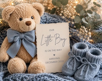 May include: A tan plush teddy bear with a light blue bow tie, a pair of light blue knitted baby booties, and a rectangular card with the text Our Little Boy IS ON THE WAY ARRIVING DECEMBER 2026, all resting on a textured blue knit fabric.