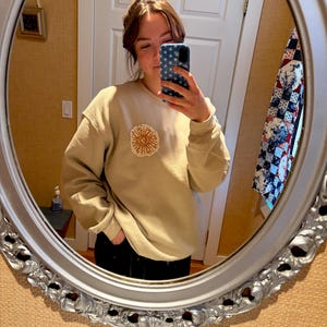 May include: A beige sweatshirt with a circular embroidered sun design on the chest. The design uses orange and beige thread. The sweatshirt has long sleeves and a relaxed fit, suitable for casual wear.
