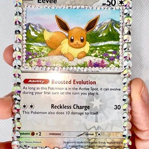 May include: A bedazzled Eevee Pokémon card with a sparkling crystal border. The card features an illustration of Eevee in a field with mountains in the background. Text includes "Eevee", "Boosted Evolution", and "Reckless Charge".