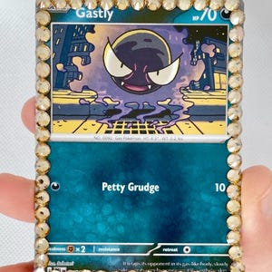 May include: A Pokémon trading card featuring Gastly, a purple ghost-type character, with a gold gem border. The card has the text "Gastly" and "Petty Grudge". The card is 4.3 inches tall and weighs 0.2 lbs.