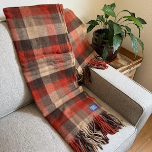 Puede incluir: Una manta a cuadros en tonos marrones, rojos y beige con bordes con flecos. La manta está sobre un sofá gris. Se ve una etiqueta azul. Un elemento de decoración acogedor para el hogar.