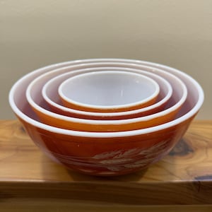 May include: Set of four nesting mixing bowls in shades of orange and white. The bowls have a wheat pattern on the exterior. The bowls are arranged in a nested configuration, with the smallest bowl at the center.