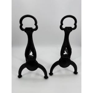 May include: A pair of black cast iron fireplace andirons featuring a tall, slender vertical post with a decorative cutout handle at the top and a three-legged base. The metal surface displays a textured, embossed pattern throughout the structure.