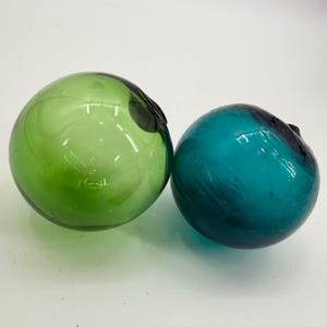 May include: Two spherical glass floats, one in a vibrant lime green and the other in a deep teal blue. Both items feature a smooth, translucent surface with a small, dark, circular indentation at the top, characteristic of hand-blown glass.