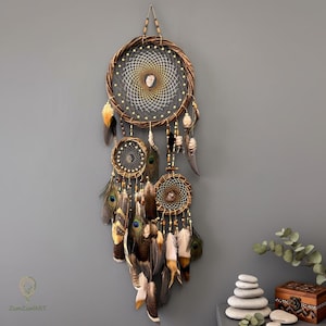 May include: A handcrafted dreamcatcher with a woven brown circular frame and intricate web design. It features multiple smaller dreamcatchers and is adorned with feathers in shades of brown, yellow, and peacock blue. The dreamcatcher is suspended against a gray wall.
