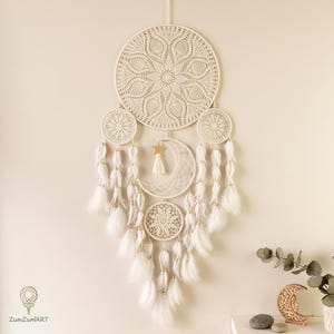 Puede incluir: Atrapasueños color crema con un gran círculo central y círculos más pequeños, una luna creciente y una estrella. Plumas blancas y cuentas doradas cuelgan de los círculos. El atrapasueños está suspendido contra una pared neutra.
