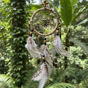 May include: A handmade dreamcatcher featuring a woven web design, decorated with white and brown feathers, wooden beads, and a small owl charm. The dreamcatcher hangs against a backdrop of green foliage.