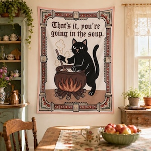 May include: A rectangular wall tapestry featuring a black cat stirring a steaming pot over a fire. The text reads, "That's it, you're going in the soup." The design is enclosed in a decorative border with pink, red, and green geometric patterns.