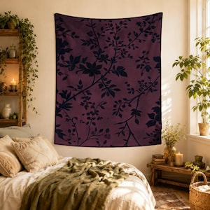 May include: A burgundy tapestry with a dark floral pattern of branches and leaves. The wall hanging is rectangular and hangs on a light-colored wall. The tapestry adds a touch of nature-inspired decor to a bedroom.