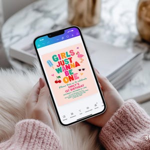 May include: A smartphone displays a pink birthday invitation with the text "GIRLS JUST WANNA BE ONE." The invitation features colorful graphics of an ice pop, cherries, sunglasses, and a flamingo. The phone is held by a person wearing a pink sweater.