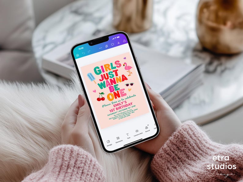 May include: A smartphone displays a pink birthday invitation with the text "GIRLS JUST WANNA BE ONE." The invitation features colorful graphics of an ice pop, cherries, sunglasses, and a flamingo. The phone is held by a person wearing a pink sweater.