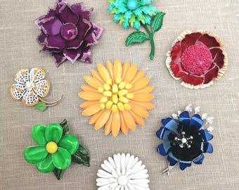 May include: A collection of vintage flower brooches in various colors, including purple, green, orange, and blue. The brooches feature different designs, such as daisies and stylized floral arrangements. Some have rhinestone accents and metal stems.