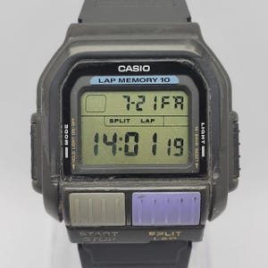 以下が含まれることがあります： 文字盤に「CASIO LAP MEMORY 10」と書かれた黒いカシオデジタル腕時計。時計は14:01:19と表示されています。長方形の画面と、紫とグレーの2つのボタンがあります。