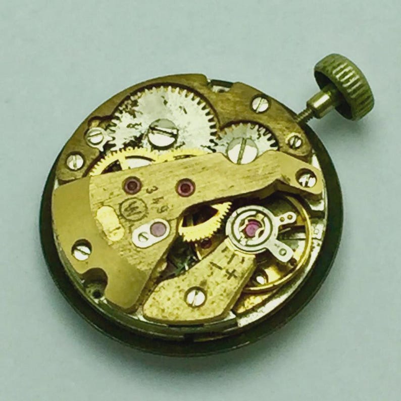 May include: Close-up of a vintage watch movement with intricate gears, jewels, and a winding stem. The metal components are gold-toned, with visible markings and a small, textured crown. The watch mechanism is circular and detailed.