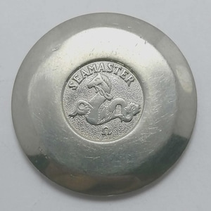 Könnte beinhalten: Silberfarbener Uhrenboden mit dem Wort "SEAMASTER" und einem Seepferdchen-Emblem in der Mitte. Der Uhrenboden ist kreisförmig mit gebürsteter Metalloberfläche und einem erhabenen zentralen Medaillon.