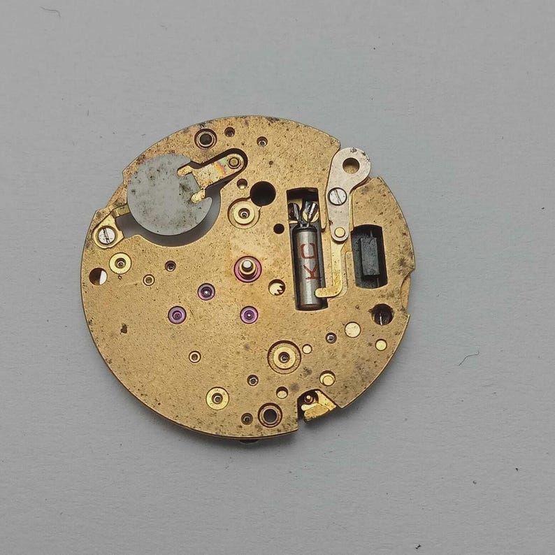 May include: Close-up of a vintage watch movement with a brass-colored base. The movement features various gears, screws, and a balance wheel. Small jewels and other components are visible, with a few components in red and purple.
