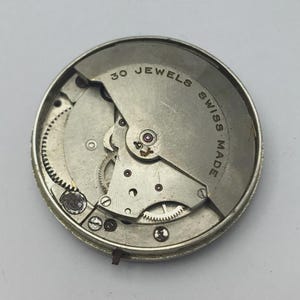K&ouml;nnte beinhalten: Nahaufnahme eines Vintage-Uhrwerks. Der silberfarbene Mechanismus zeigt komplizierte Zahnr&auml;der, eine Unruh und den Text "30 JEWELS SWISS MADE". Die Uhrwerkkomponenten sind in einem kreisf&ouml;rmigen Rahmen untergebracht.