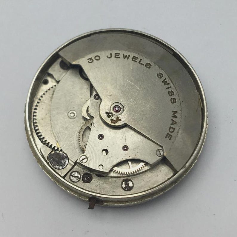 K&ouml;nnte beinhalten: Nahaufnahme eines Vintage-Uhrwerks. Der silberfarbene Mechanismus zeigt komplizierte Zahnr&auml;der, eine Unruh und den Text "30 JEWELS SWISS MADE". Die Uhrwerkkomponenten sind in einem kreisf&ouml;rmigen Rahmen untergebracht.