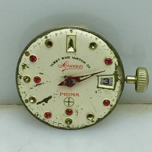 May include: Vintage watch face with a cream-colored dial. The face features the text "WEST END WATCH Co Sowar PRIMA" in red and black. It has red and clear gem accents, a date window, and gold-colored hands and crown.