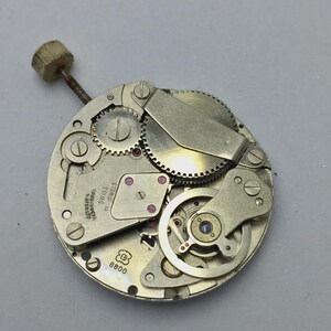 Pu&ograve; includere: Primo piano di un movimento di orologio con ingranaggi, viti e una corona visibili. Il movimento &egrave; argentato con gioielli rossi e la scritta "SWISS 17 JEWELS". Un componente di orologio vintage.