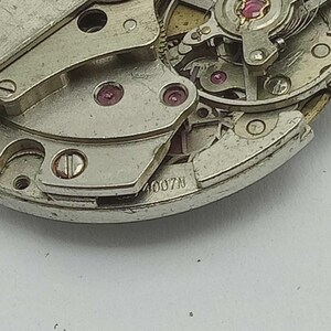 May include: Detailed view of a watch mechanism, showcasing silver-toned metal components, tiny red jewels, and the text "4007N". The image focuses on the intricate gears and precision engineering within the watch's movement.