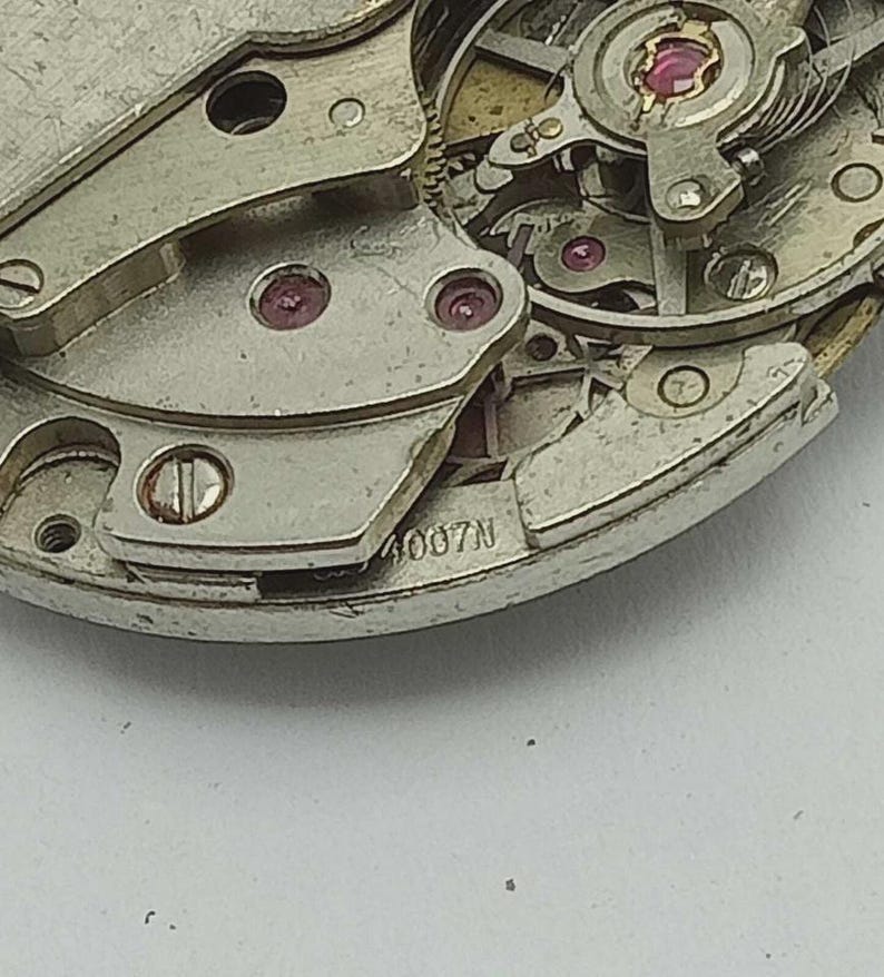 May include: Detailed view of a watch mechanism, showcasing silver-toned metal components, tiny red jewels, and the text "4007N". The image focuses on the intricate gears and precision engineering within the watch's movement.