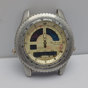 Op de afbeelding: Een vintage Casio Quartz Yacht timer horloge met een zilverkleurige kast en een crèmekleurige wijzerplaat. Het horloge heeft een draaibare bezel, meerdere wijzerplaten en de tekst "Yacht timer 100M". Het horloge is gebruikt.