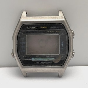 May include: A silver Casio digital watch case with a black display. The watch face includes the text "CASIO", "Lithium", "Water Resist 10CM", and "Alarm Chronograph". The watch has buttons on the sides and a rectangular display.