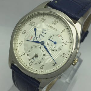 May include: Silver-toned J. Springs watch with a white face, blue hands, and a blue leather band. The watch face displays the day, date, and seconds. The watch has a round face with Arabic numerals and a textured design.