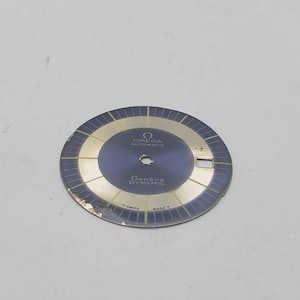 May include: A vintage Omega Genève Dynamic watch dial. The dial features a dark blue center with gold-colored accents and text. The text includes "OMEGA AUTOMATIC" and "Genève DYNAMIC". The dial has a circular shape.