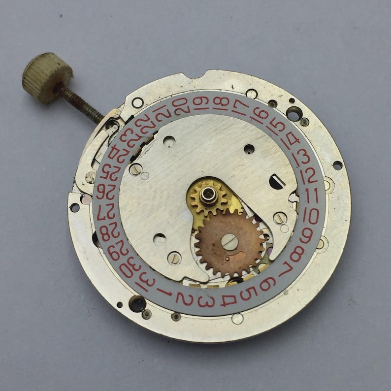 Pu&ograve; includere: Primo piano di un movimento di orologio con una base in metallo color argento. Il movimento presenta un datario con numeri rossi, un piccolo ingranaggio dorato e una vite con una testa color beige. Il meccanismo dell'orologio &egrave; un esempio dettagliato di orologeria.