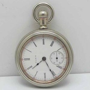 May include: A silver Elgin pocket watch with a white face and black Roman numerals. The watch has a small seconds dial and a textured crown. The watch is round and has a silver bezel.