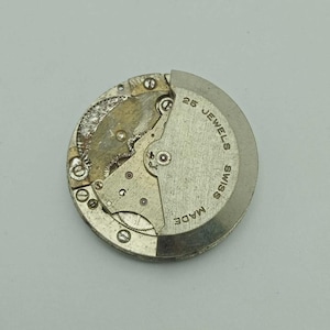 May include: A detailed view of a watch movement, showcasing its internal mechanisms. The metal components are silver and gold in colour, with the text "25 JEWELS SWISS MADE" inscribed on the circular base. A vintage watch mechanism.