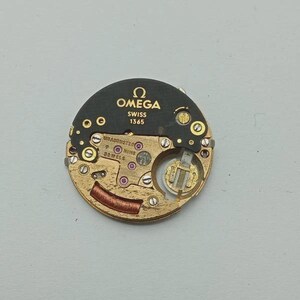 May include: A close-up of an Omega watch movement, featuring a gold-toned base with intricate gears, jewels, and a copper-colored coil. The black upper portion displays the Omega logo, "SWISS 1365," and other markings. The watch movement is a precision mechanical component.
