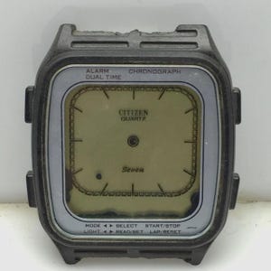 Puede incluir: Una esfera de reloj digital Citizen Quartz vintage con una caja negra y un bisel plateado. La esfera del reloj muestra el texto "ALARM DUAL TIME CHRONOGRAPH" y "CITIZEN QUARTZ". El reloj tiene varios botones de función y la palabra "Seven" es visible.